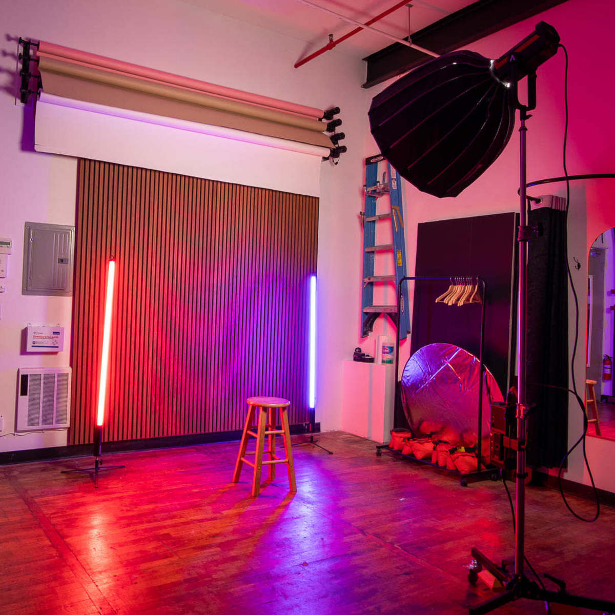 A photography studio features a stool in the center, flanked by neon lights, with a large softbox and reflective surfaces visible in the background.