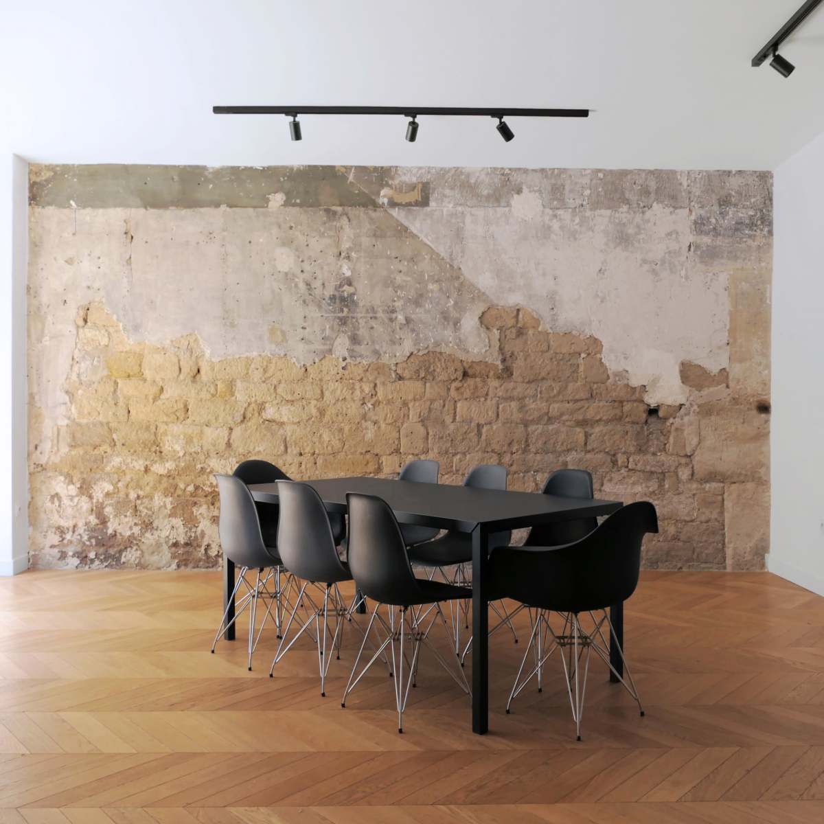 A modern dining area features a large black table surrounded by sleek black chairs, set against a rustic textured wall and wooden herringbone flooring.