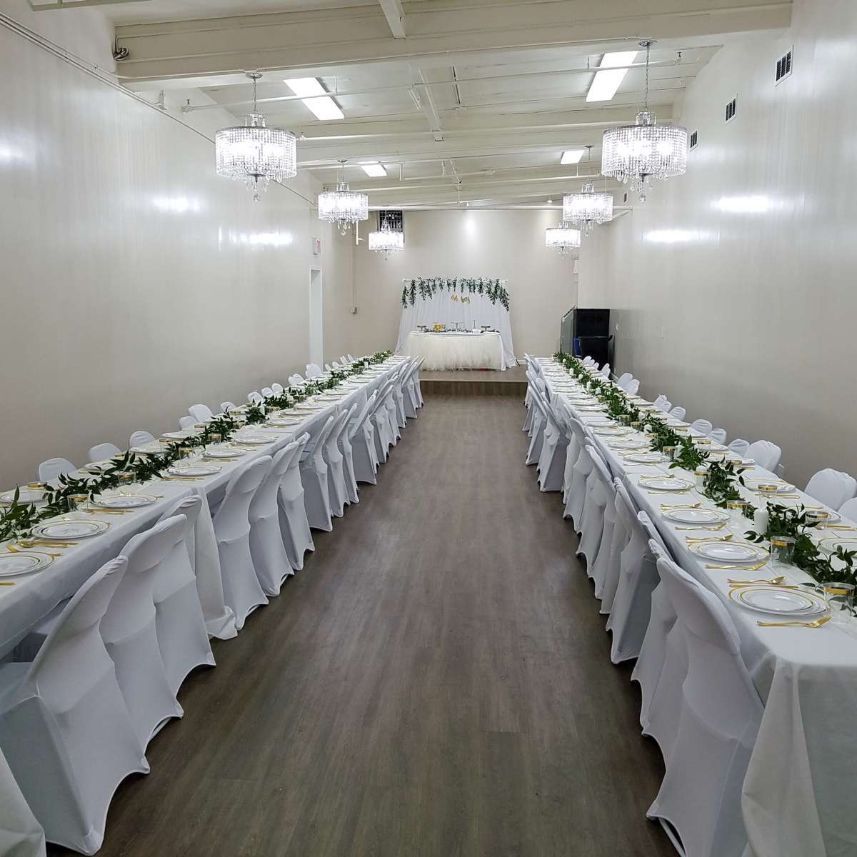 A long banquet hall with two rows of elegantly set tables featuring white tablecloths and greenery decorations, leading to a decorated stage at the far end.