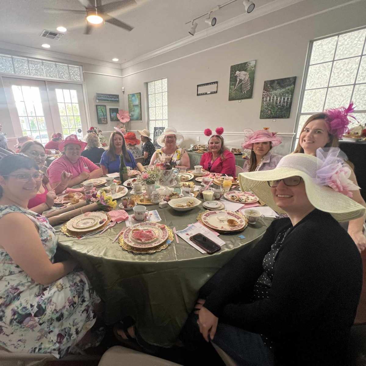 A group of women wearing large hats gathers around a table set for a tea party, with various dishes and decorative items in a well-lit room.