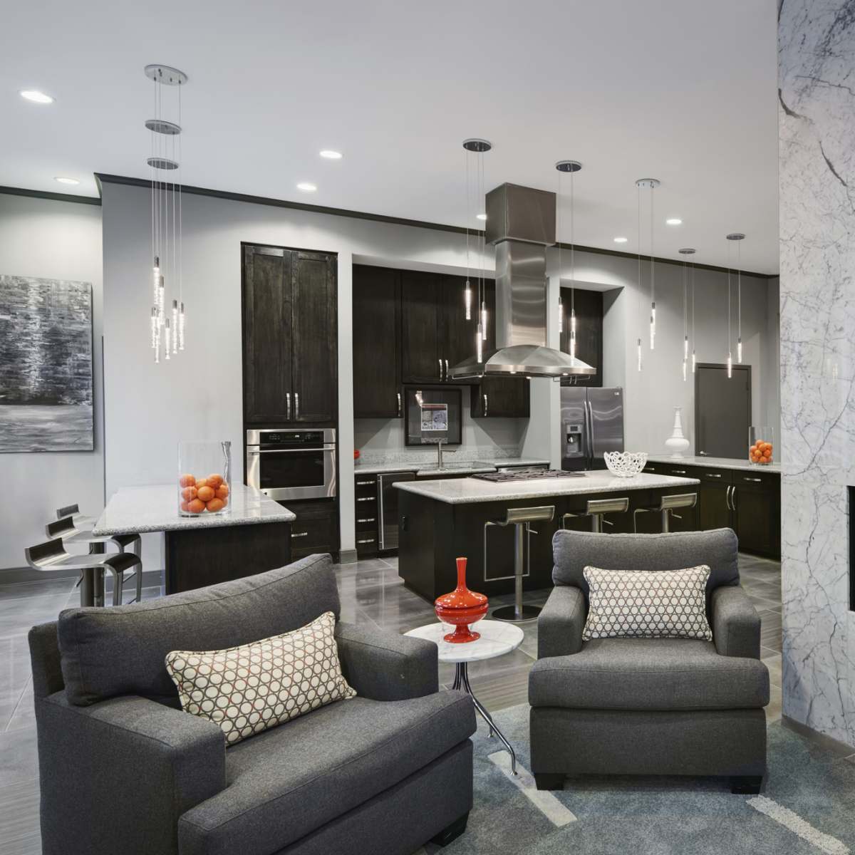 A modern kitchen and living area features gray armchairs, a marble fireplace, and sleek cabinetry with overhead lighting.