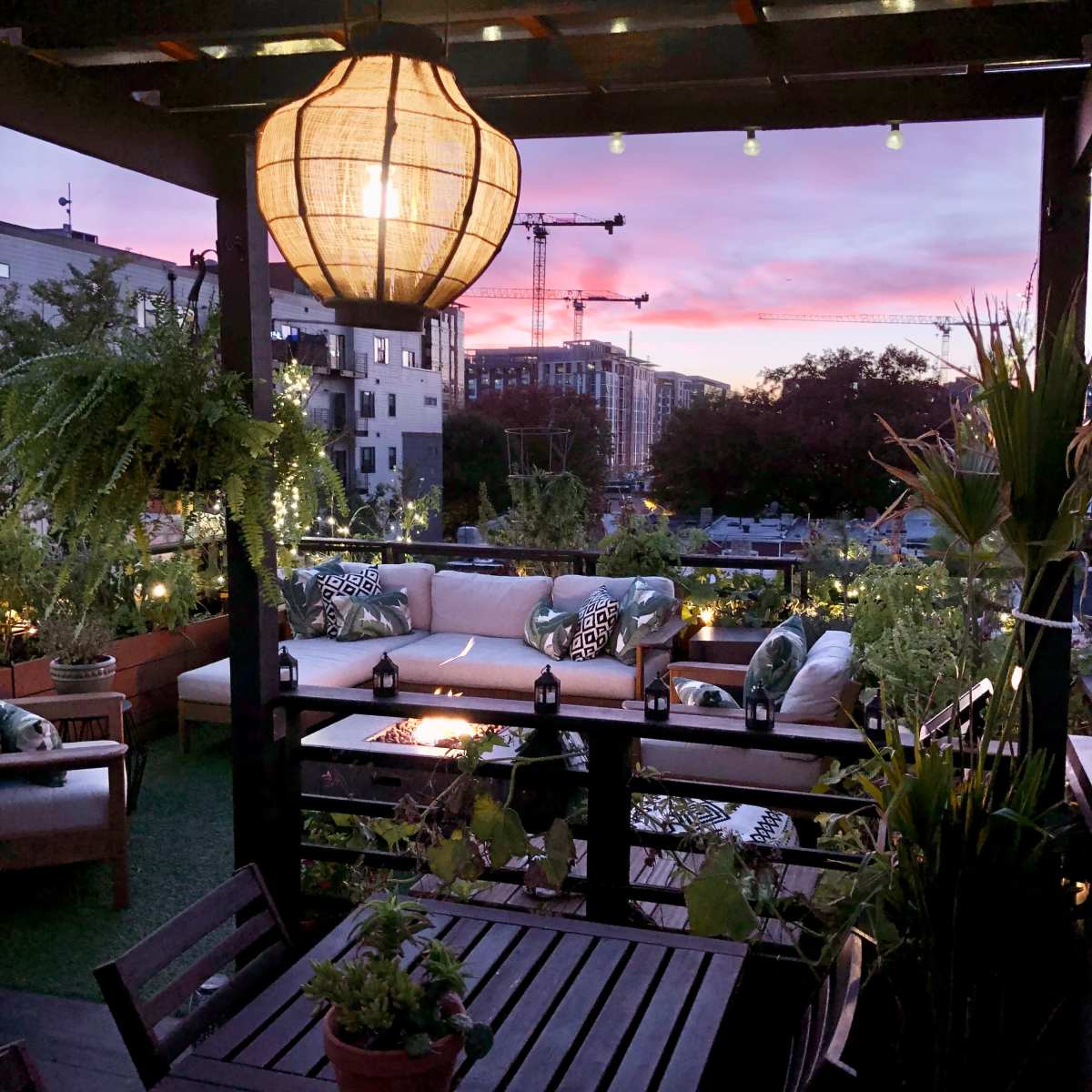 A rooftop terrace features a lantern, comfortable seating, and potted plants under a colorful sunset sky.