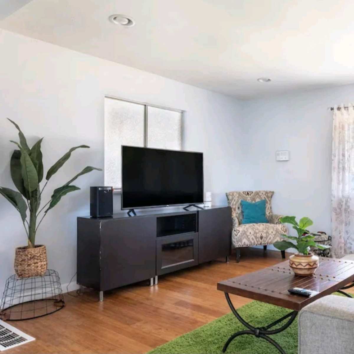 A modern living room features a television on a black console, a light-colored sofa, a wooden coffee table, and decorative plants.