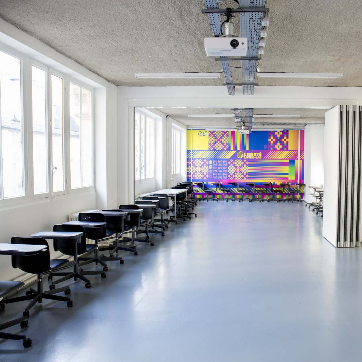 A spacious classroom with rows of black wheeled chairs and a colorful geometric mural on one of the walls.