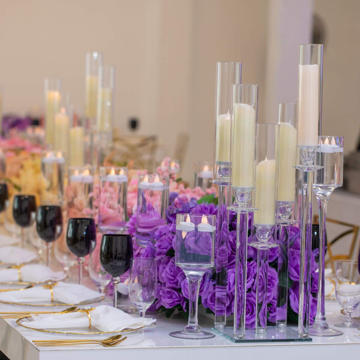 A banquet table is set with white plates, gold flatware, and an arrangement of purple roses alongside towering candles and decorative glassware.