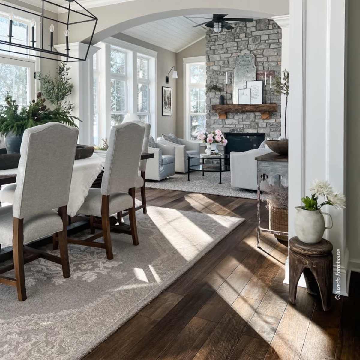 A bright, inviting living space featuring a dining area with upholstered chairs, a stone fireplace, and large windows allowing natural light to fill the room.
