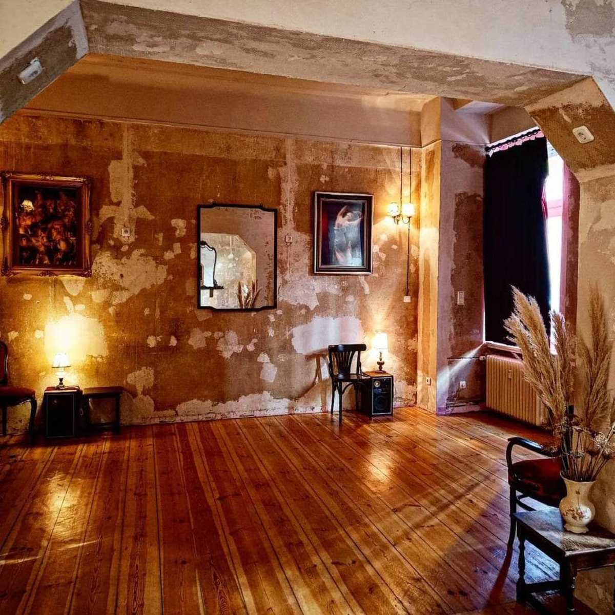 A sparsely furnished room with wooden flooring, featuring a bare wall, two light sources, and a few decorative items, including a vase with dried plants.