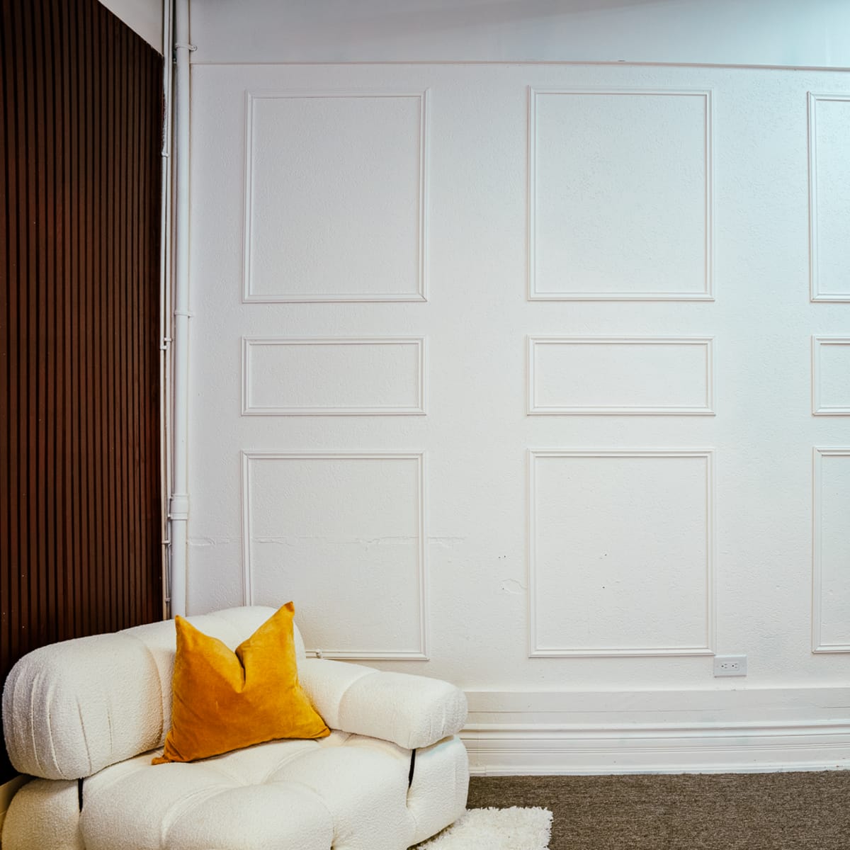 A corner of a room featuring a textured white wall with decorative moldings, a light-colored couch with an orange pillow, and a white door.