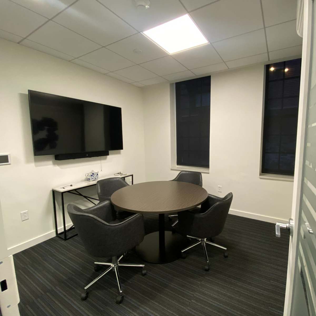A small conference room with a round table, four chairs, a wall-mounted television, and large windows.