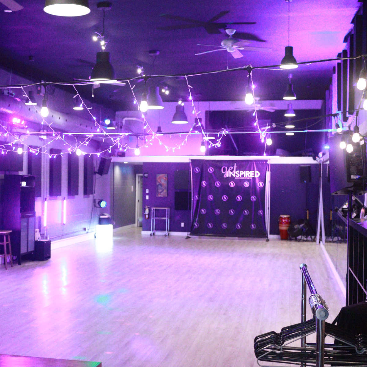 A spacious dance studio with purple lighting, wooden flooring, and string lights overhead.