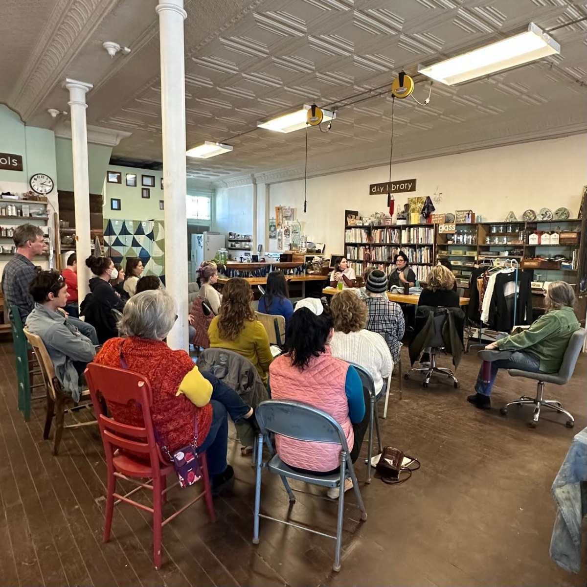 A group of people sits in chairs, facing a table with speakers, in a spacious, well-lit community room filled with bookshelves and decor.
