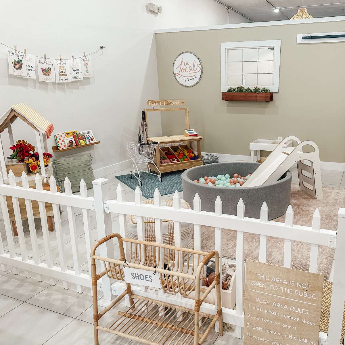 A children's play area features a sandbox, slide, playhouse, and toy storage, enclosed by a white picket fence.