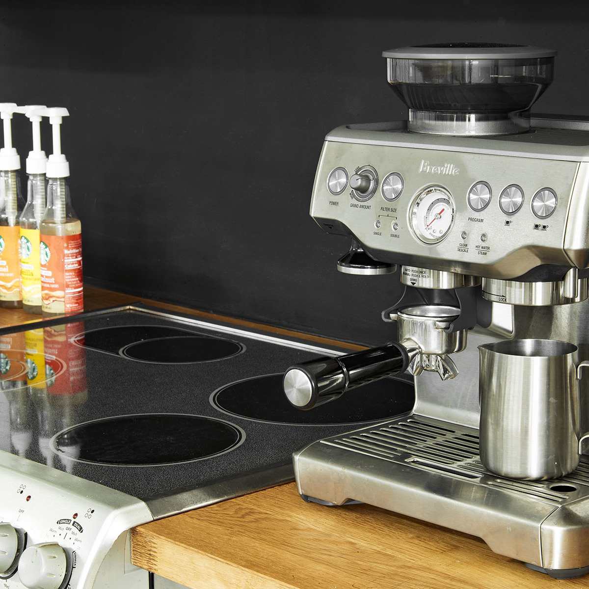 A stainless steel espresso machine is positioned on a wooden countertop beside a smooth cooktop.