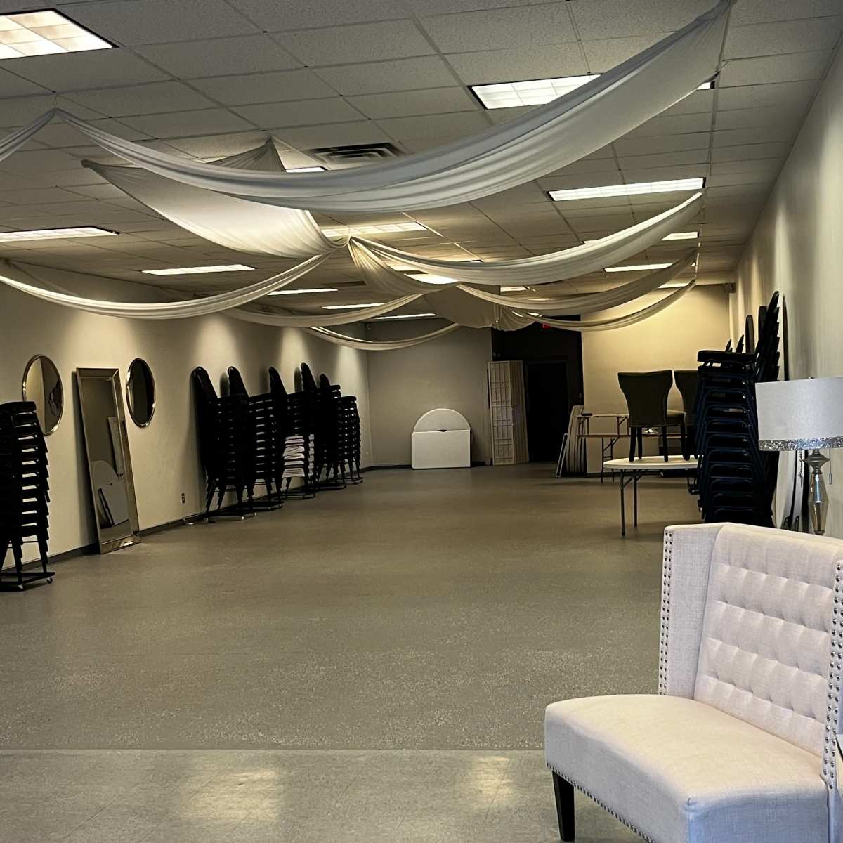 An empty event space with fabric draped from the ceiling, stacked chairs against the wall, and a decorative chair in the foreground.