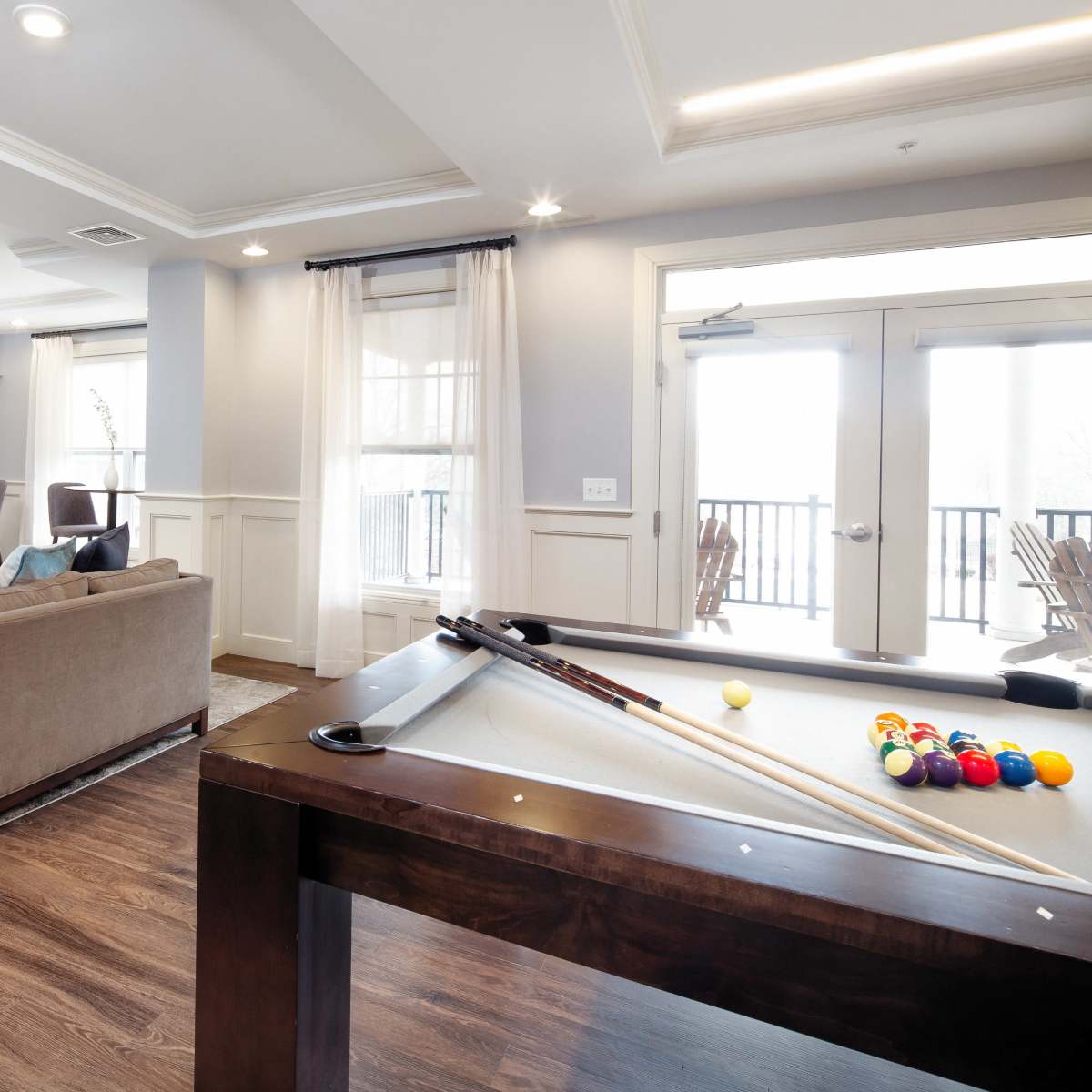 A modern living room with a pool table, a cozy sofa facing a television mounted above a fireplace, and large windows leading to a balcony.