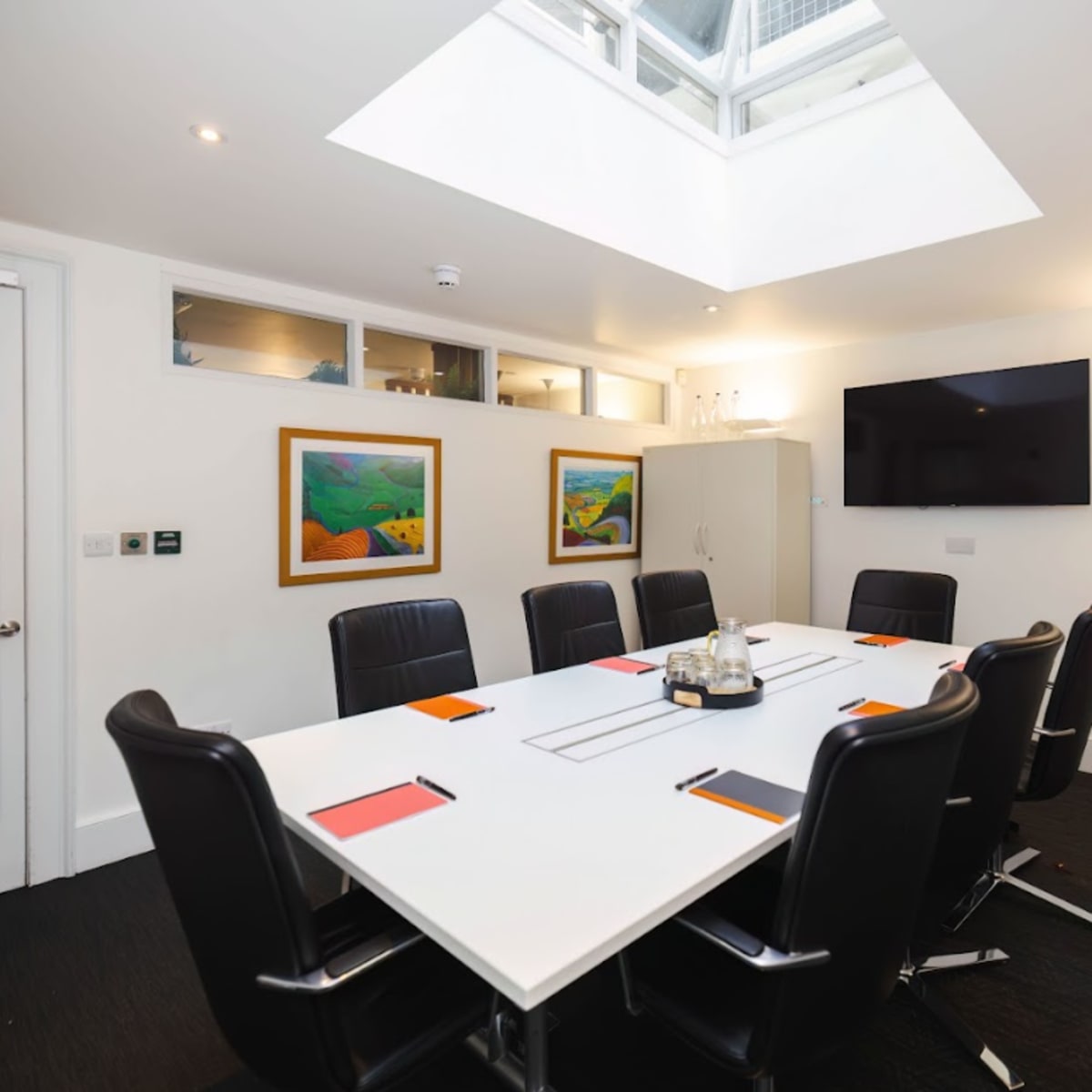 A modern conference room features a large white table surrounded by black chairs, with paintings on the walls and a skylight above.