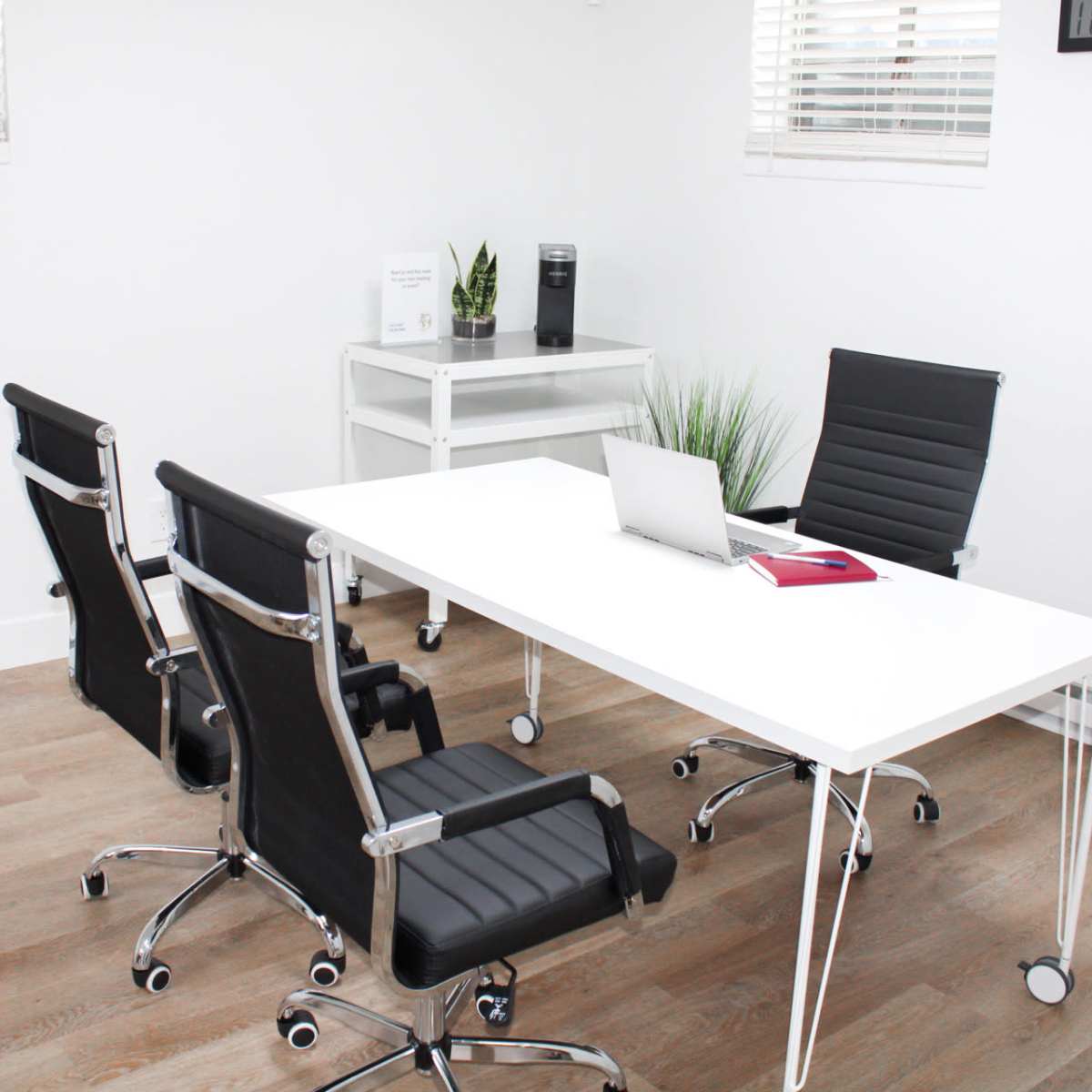 A modern office space features a white table, two black ergonomic chairs, a laptop, and potted plants against white walls.
