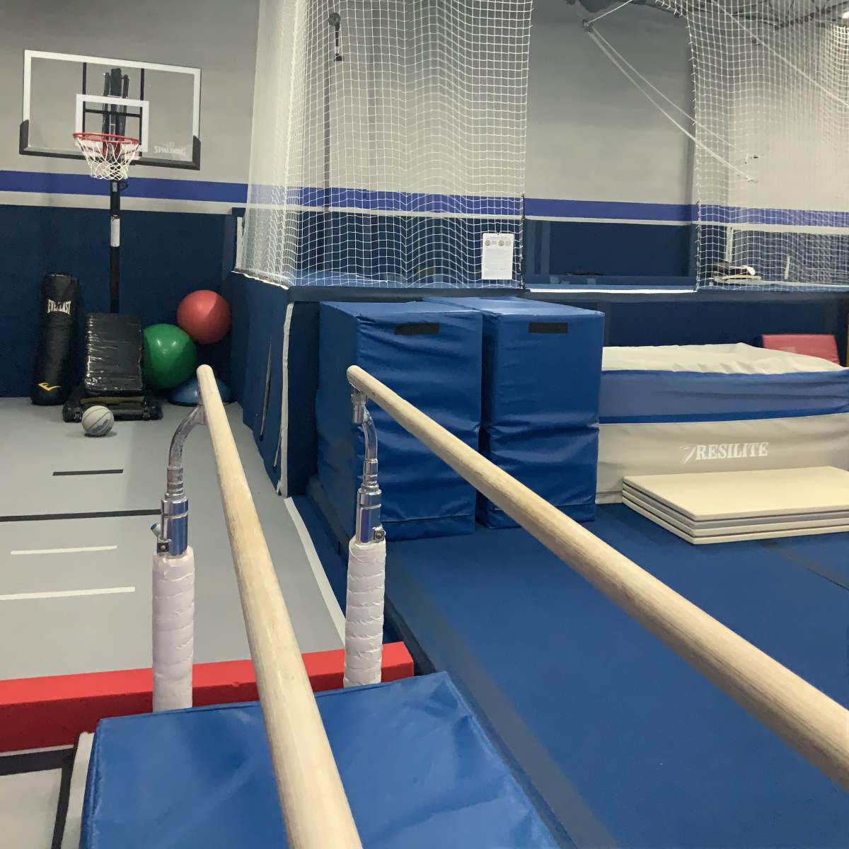 A gymnastics training facility with parallel bars in the foreground, gym mats, and a basketball hoop in the background.