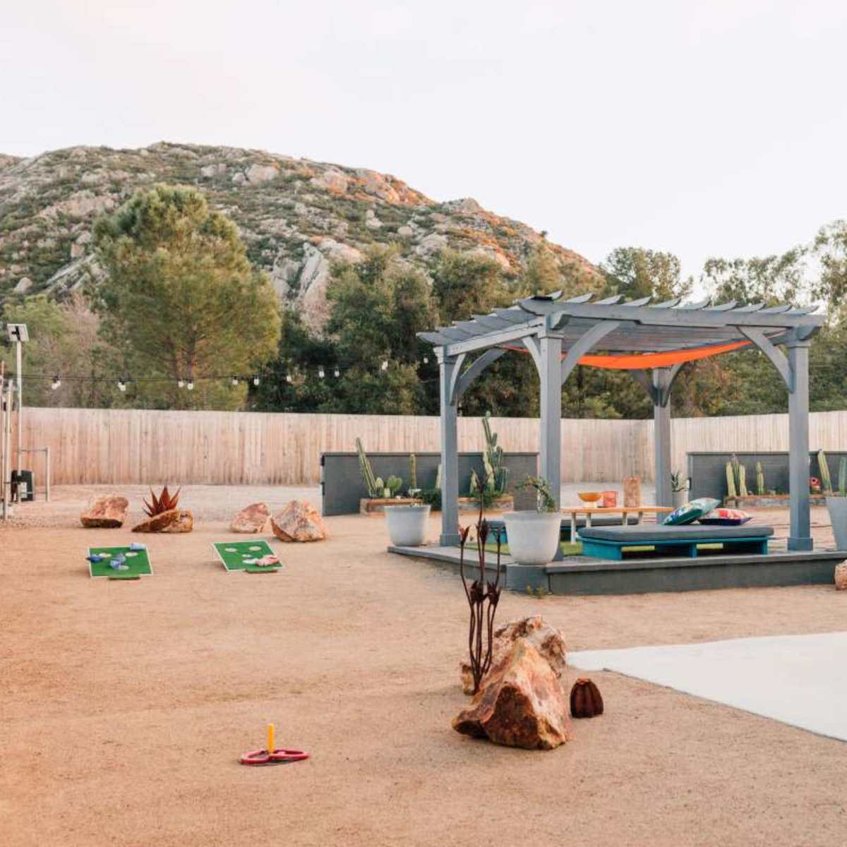 A landscaped outdoor area featuring a mini-golf setup, a wooden pergola with seating, and rock formations against a backdrop of trees and mountains.
