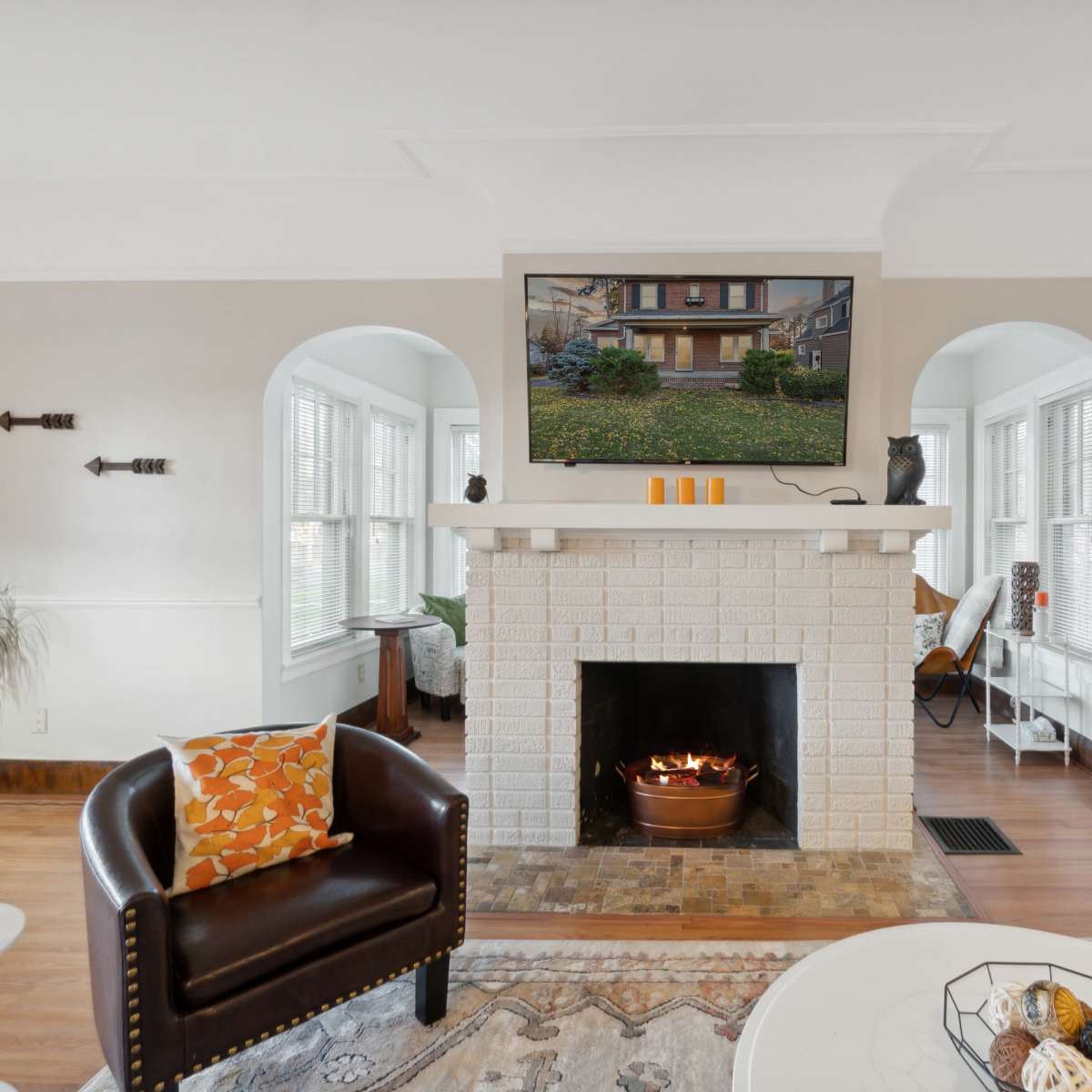 A cozy living room featuring a white brick fireplace with a mounted screen above, a brown leather armchair, and large windows allowing natural light to fill the space.