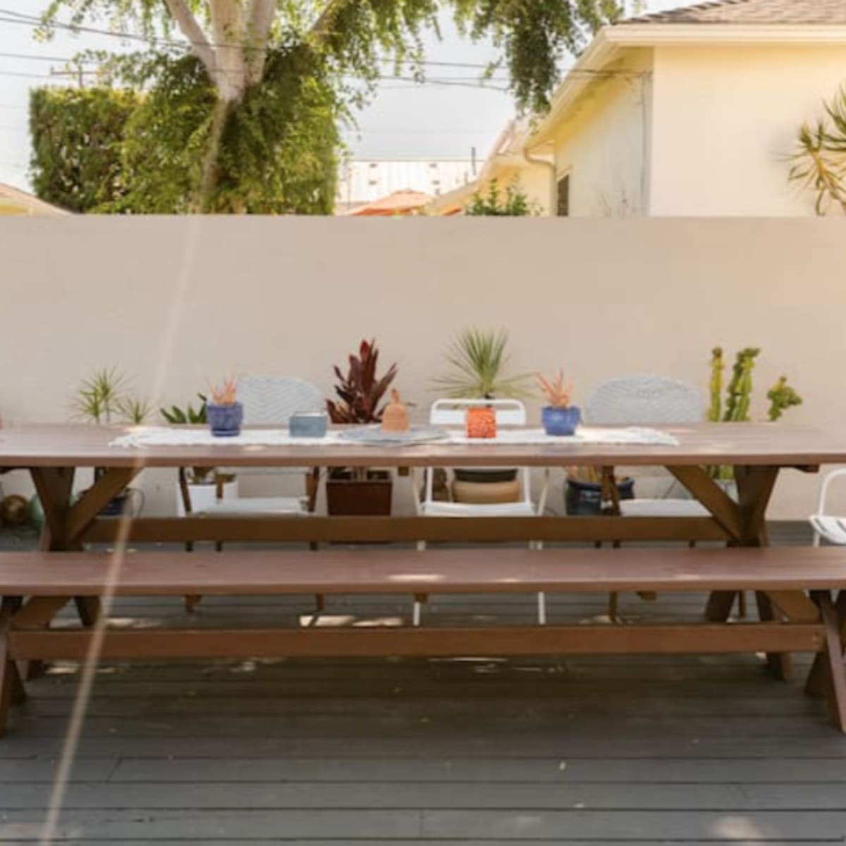 A spacious wooden dining table set in a minimalistic outdoor area with potted plants and two white chairs on either side.