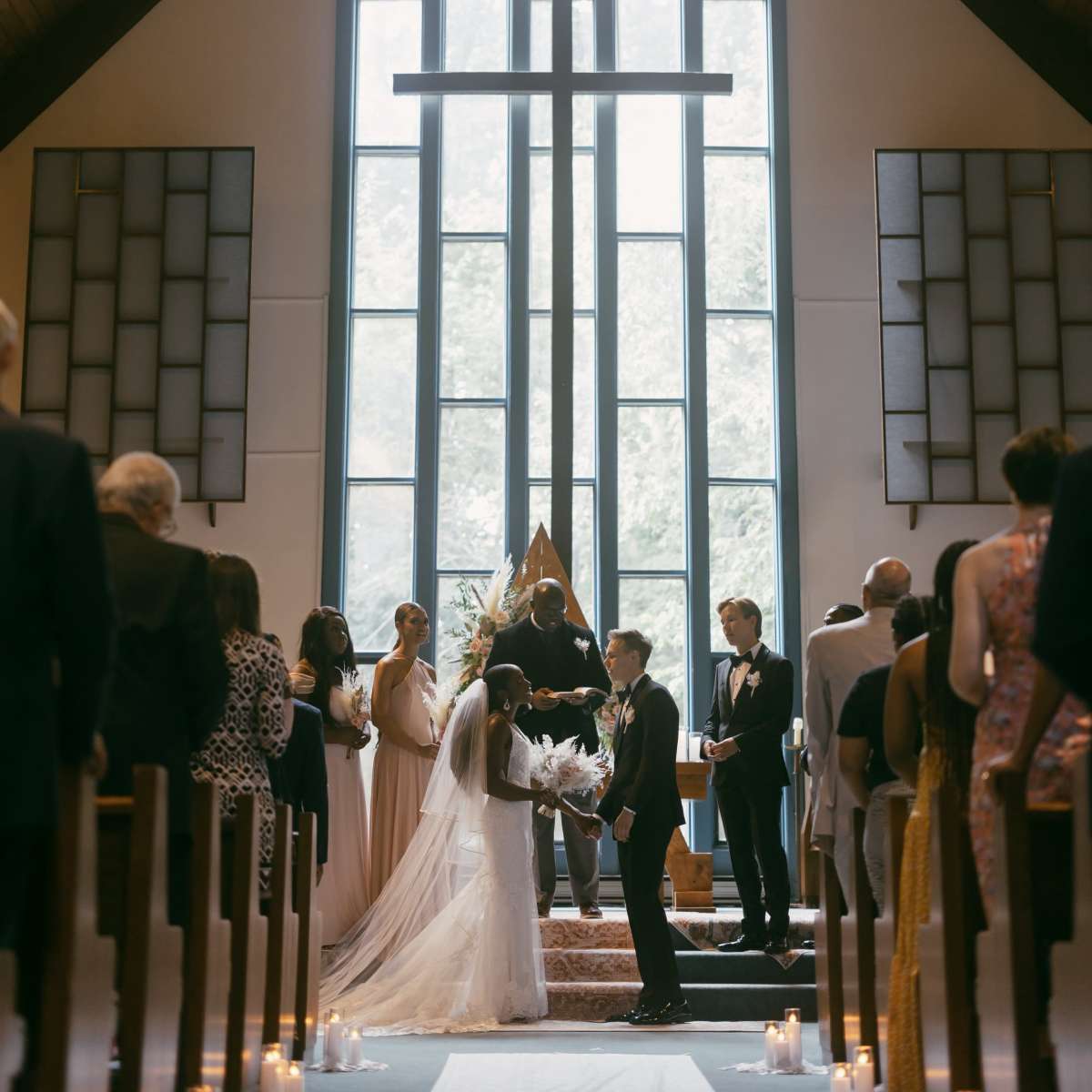 A bride and groom stand under a large cross during their wedding ceremony in a church, surrounded by guests and bridal party members.