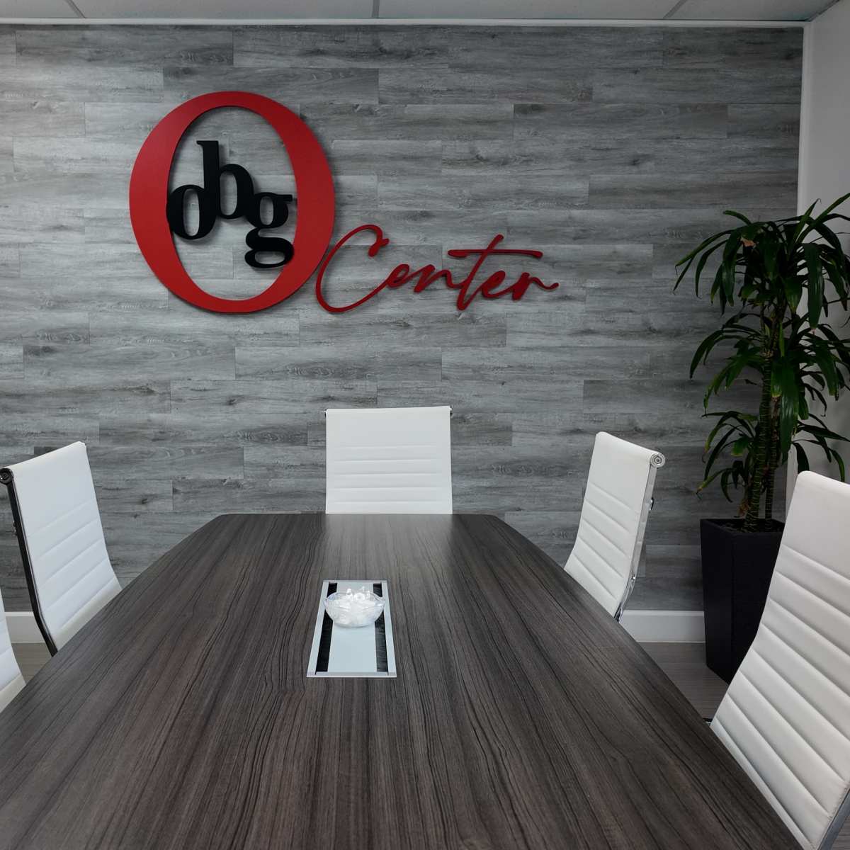 A conference room features a long table surrounded by white chairs, with a large red and black logo on the wall.