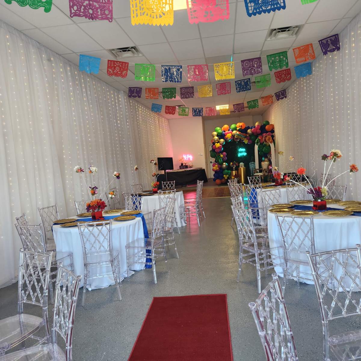 A decorated event space with tables covered in white tablecloths, surrounded by transparent chairs, and colorful papel picado banners hanging from the ceiling.