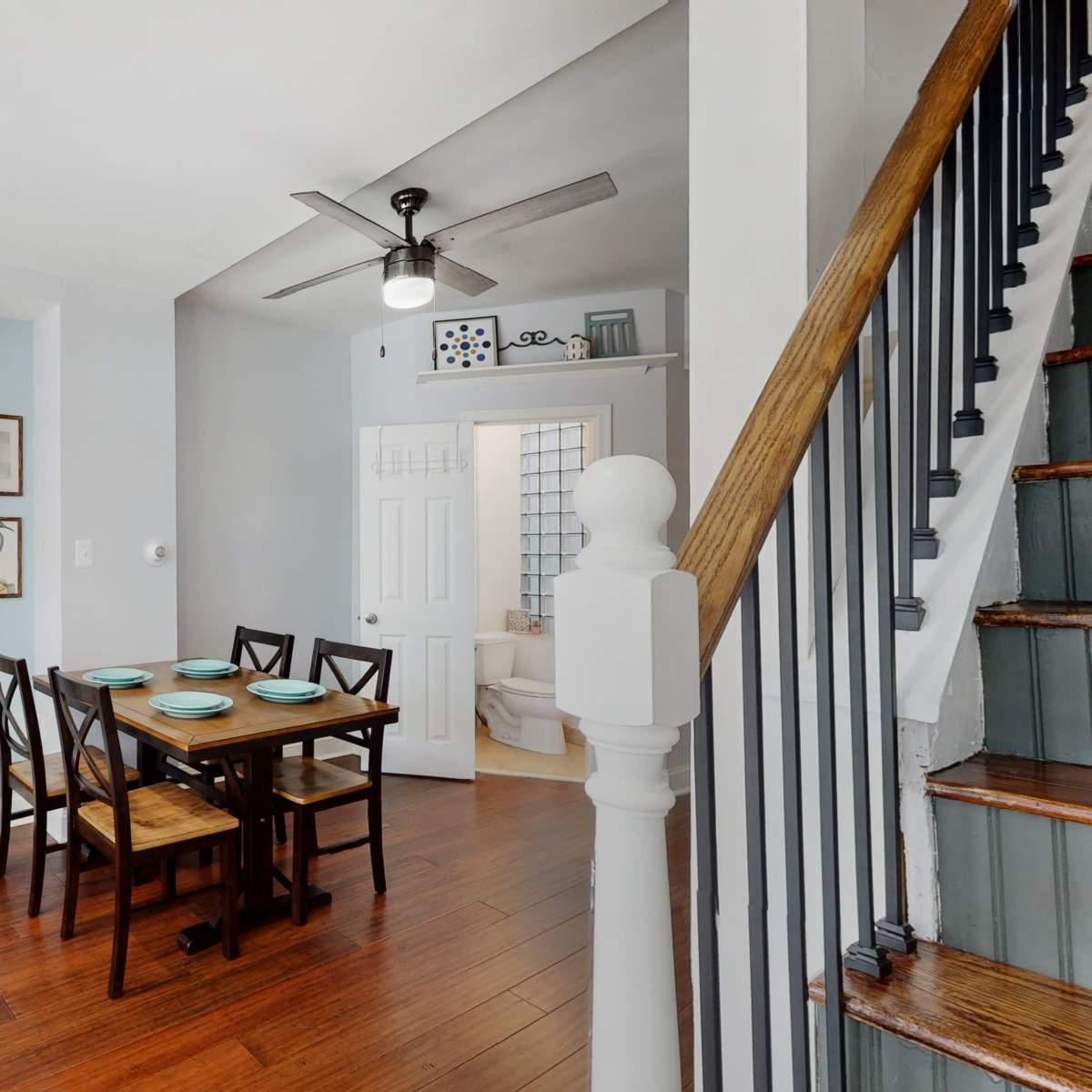 A well-lit dining area featuring a table with four chairs, surrounded by light blue walls and hardwood flooring, with a staircase visible in the background.
