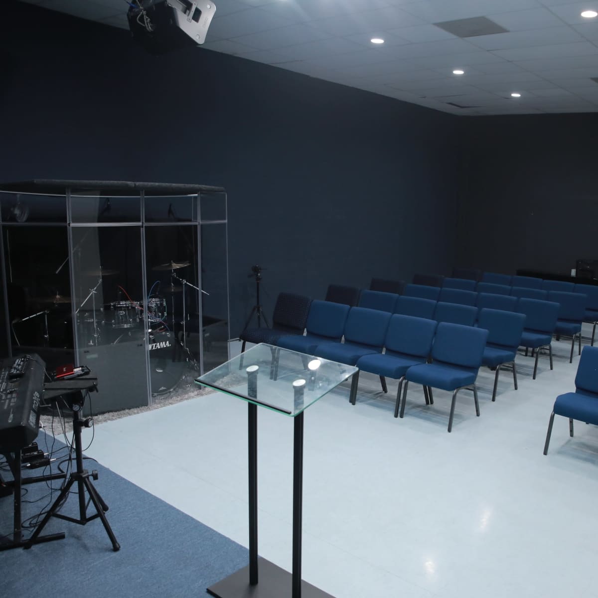 An empty room features blue chairs arranged in rows facing a glass-enclosed drum set and a transparent lectern.