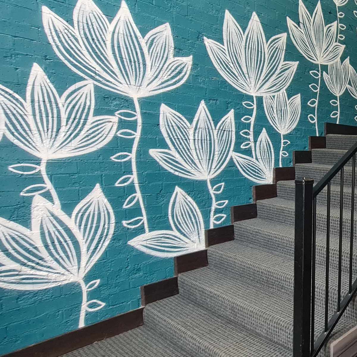 A stairwell with a teal mural featuring white floral designs leads to a landing with informational posters and a bench.