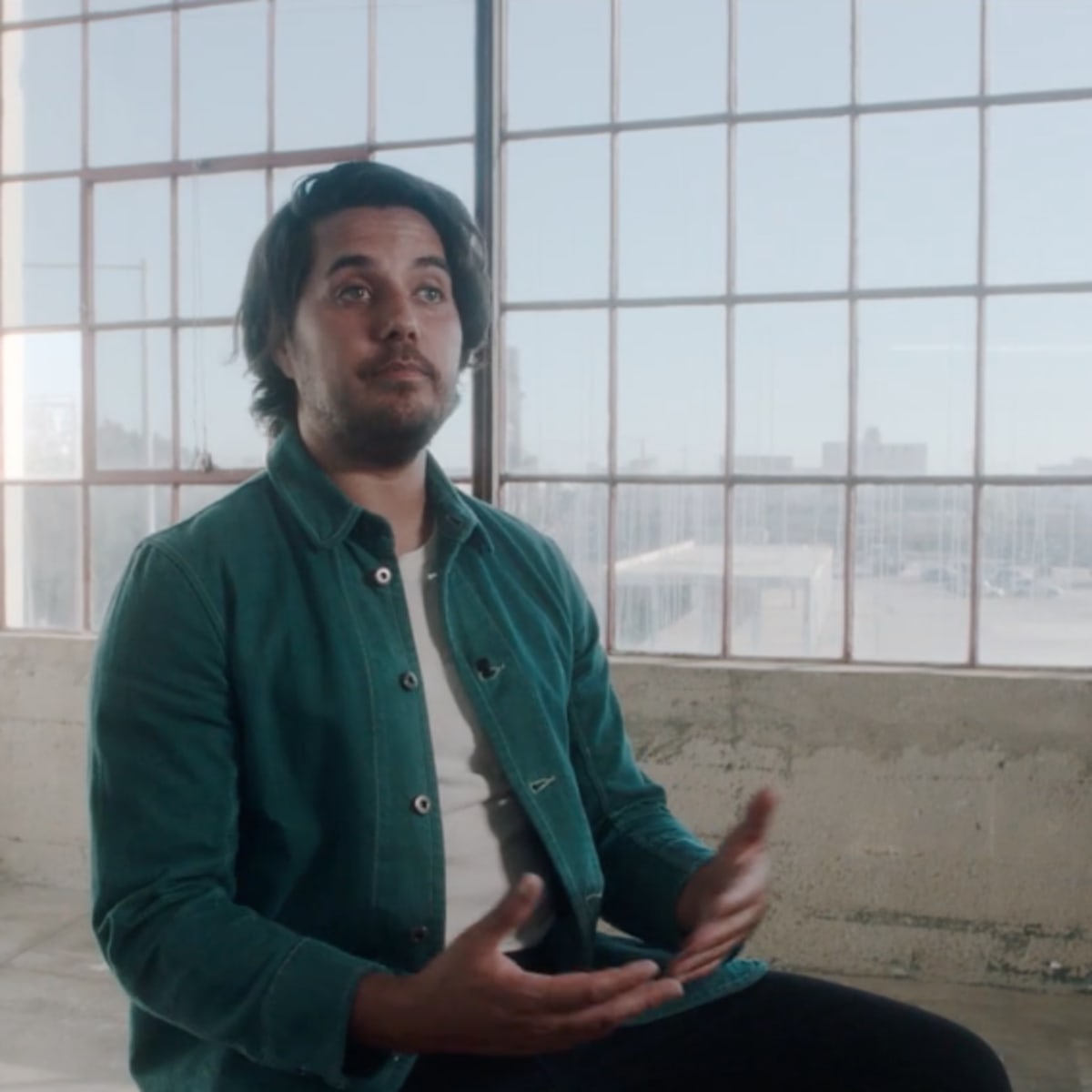 A man sits with his hands gesturing in a sunlit industrial space with large windows.