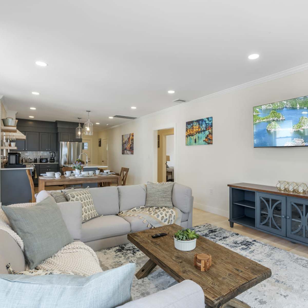 The living room features a light-colored sofa, a wooden coffee table, and a wall-mounted television, with a dining area visible in the background.