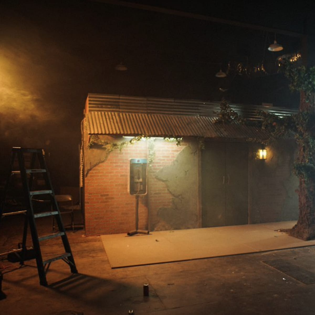 A staged interior set with a brick wall, a tree, a payphone, and various lighting equipment in a dimly lit space.
