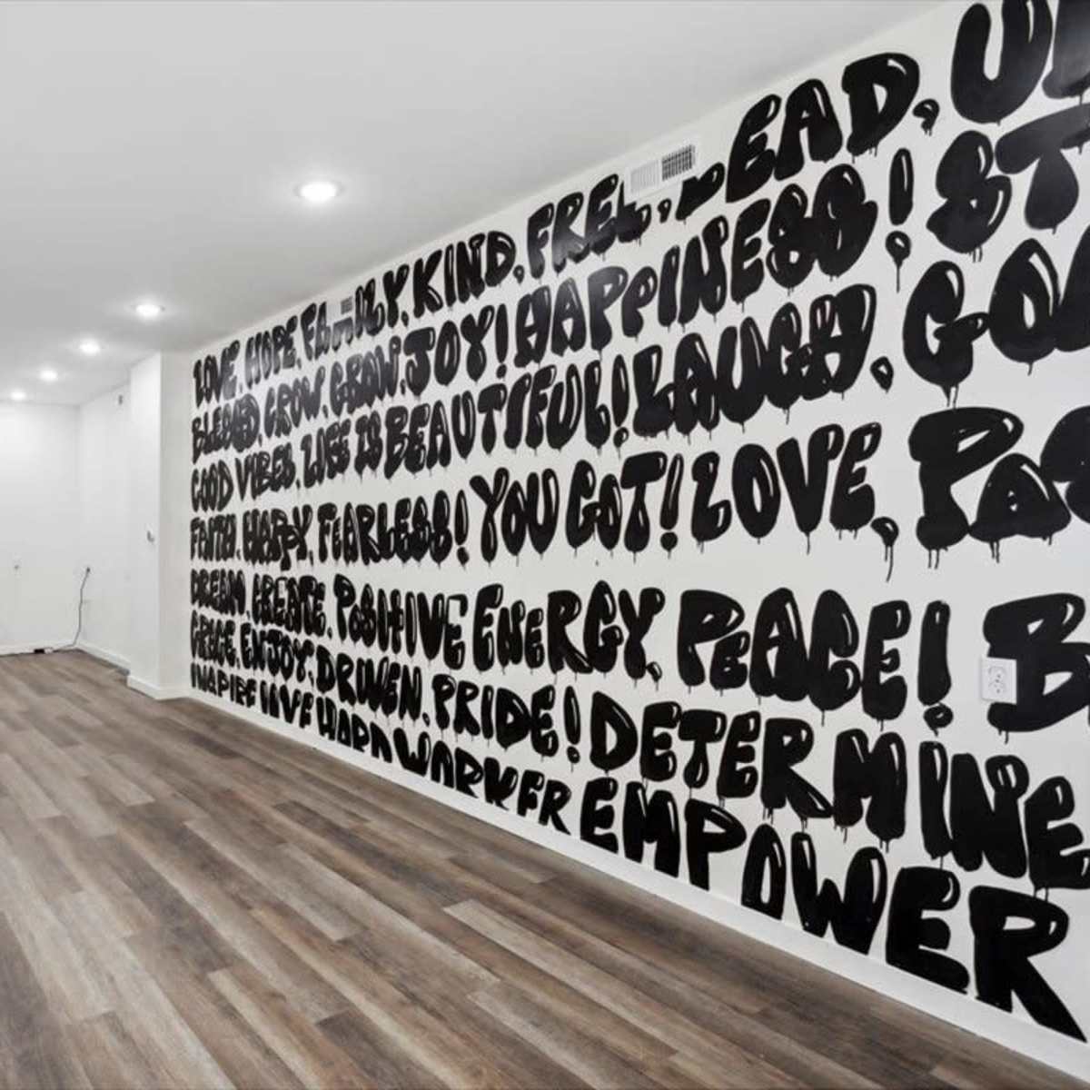 A spacious, empty room with wooden flooring and a wall covered in large, bold black text, while two chairs are positioned against the opposite wall.