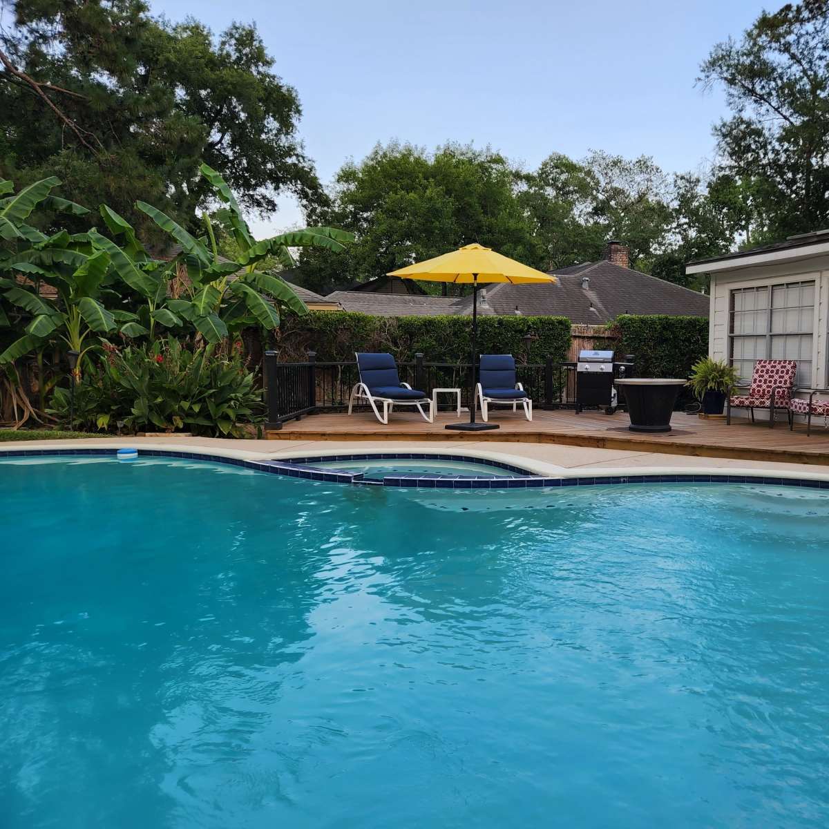 A swimming pool surrounded by sun loungers, an umbrella, and several potted plants next to a house.
