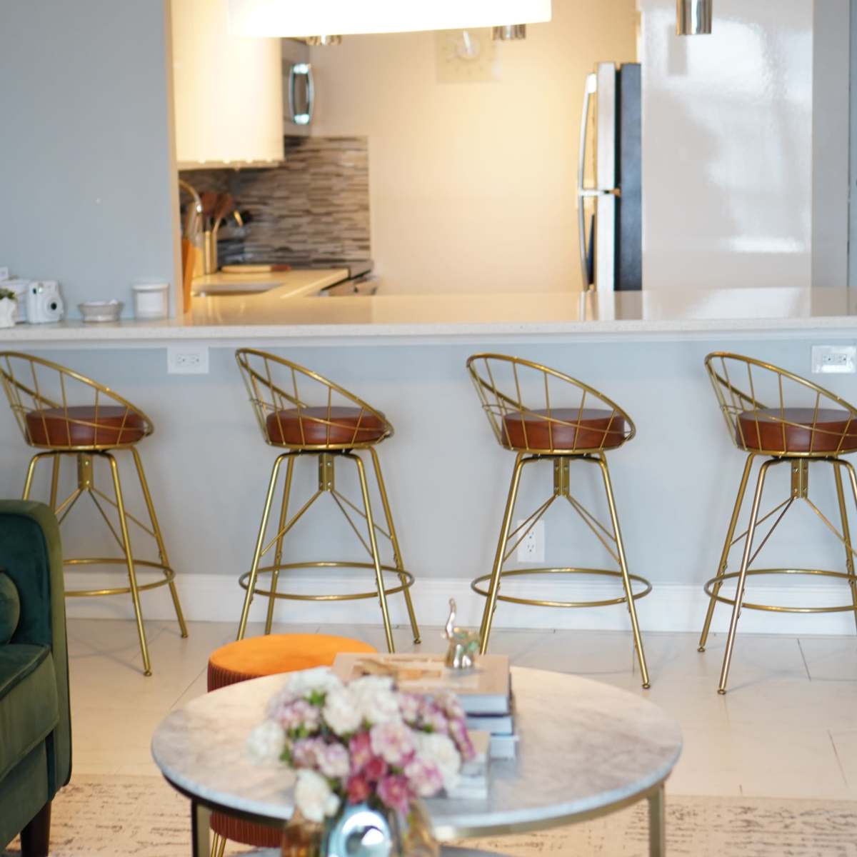 A modern kitchen bar area features four gold-accented stools lined up at a white countertop, with a glimpse of kitchen appliances and decor in the background.