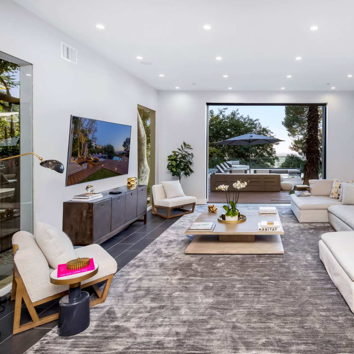 A contemporary living room with large windows, a light-colored sectional sofa, and a flat-screen TV mounted on the wall.