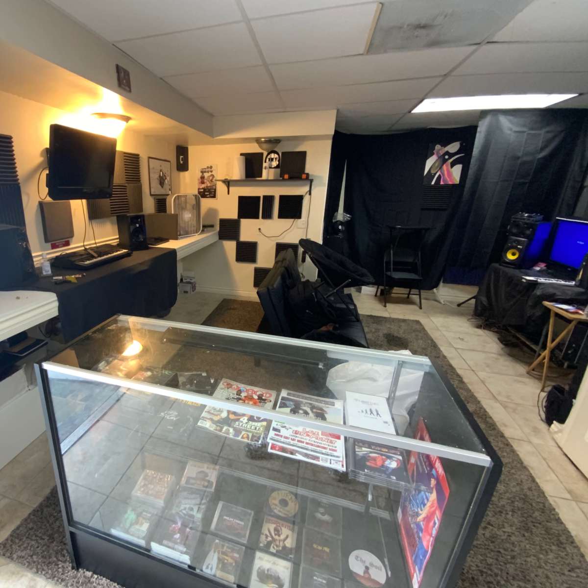 A room with a glass display case filled with DVDs, a desk with electronic devices, and black fabric draped across the walls, creating a studio-like atmosphere.