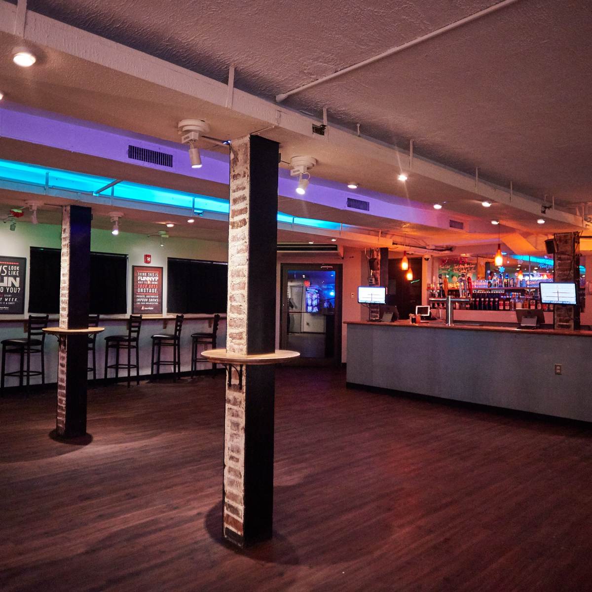 An empty bar interior features a counter with colorful lighting, exposed brick pillars, and seating arranged against the walls.