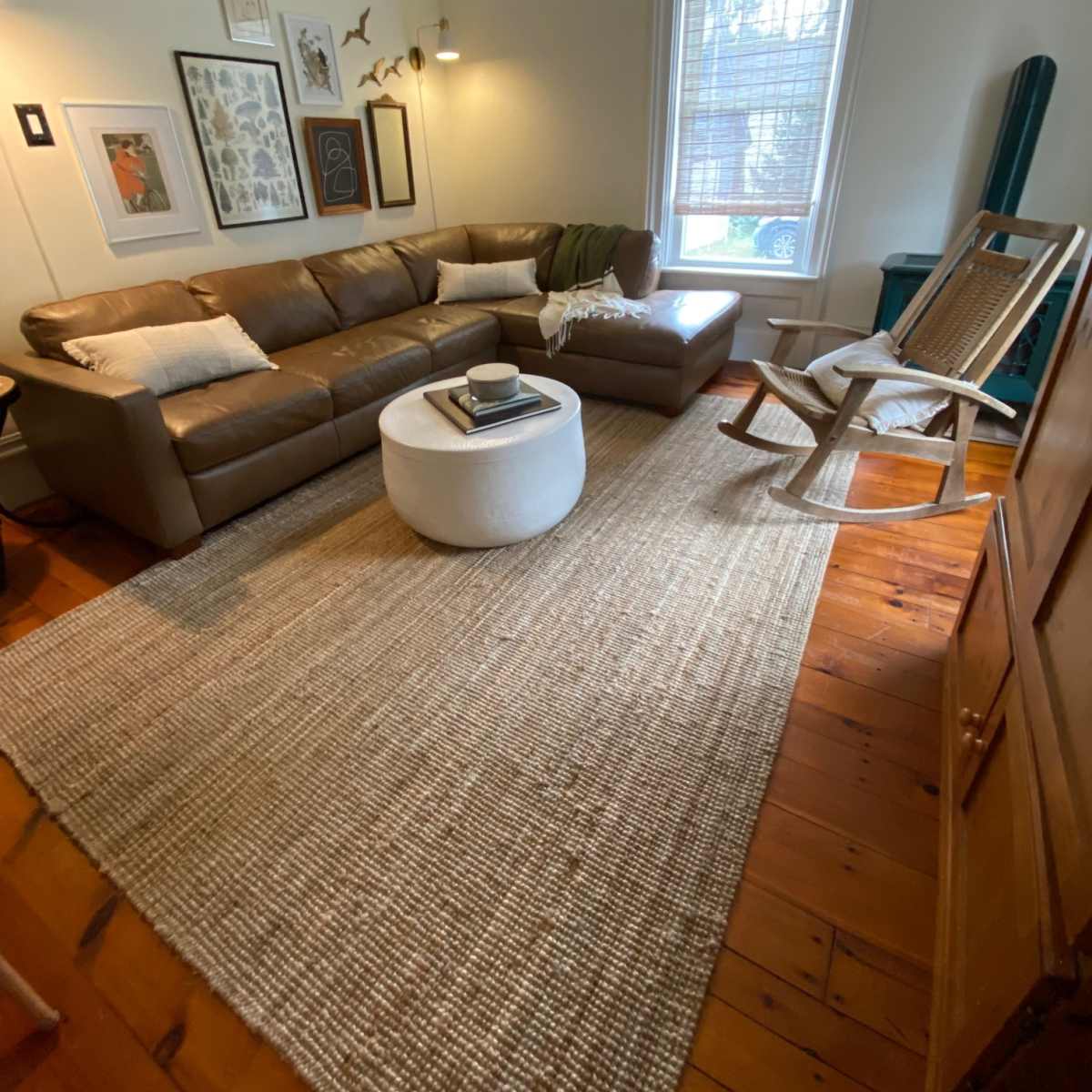 A living room with a brown sectional sofa, a round coffee table, a rocking chair, and a large area rug on wooden flooring.
