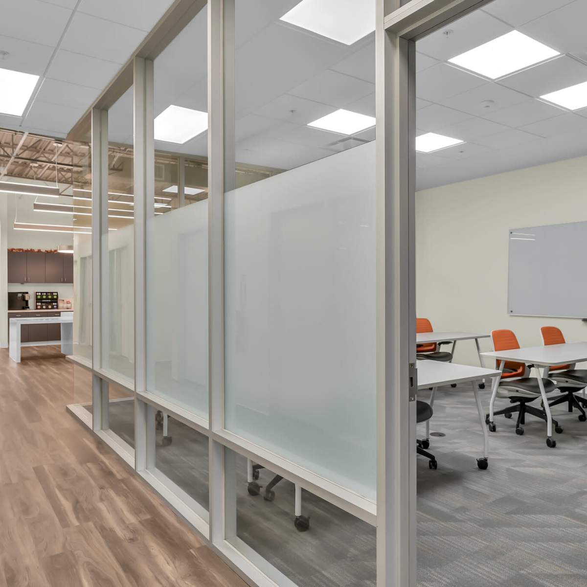 A modern office space with glass-paneled meeting rooms, orange chairs, and a hallway leading to a well-lit area with a whiteboard.