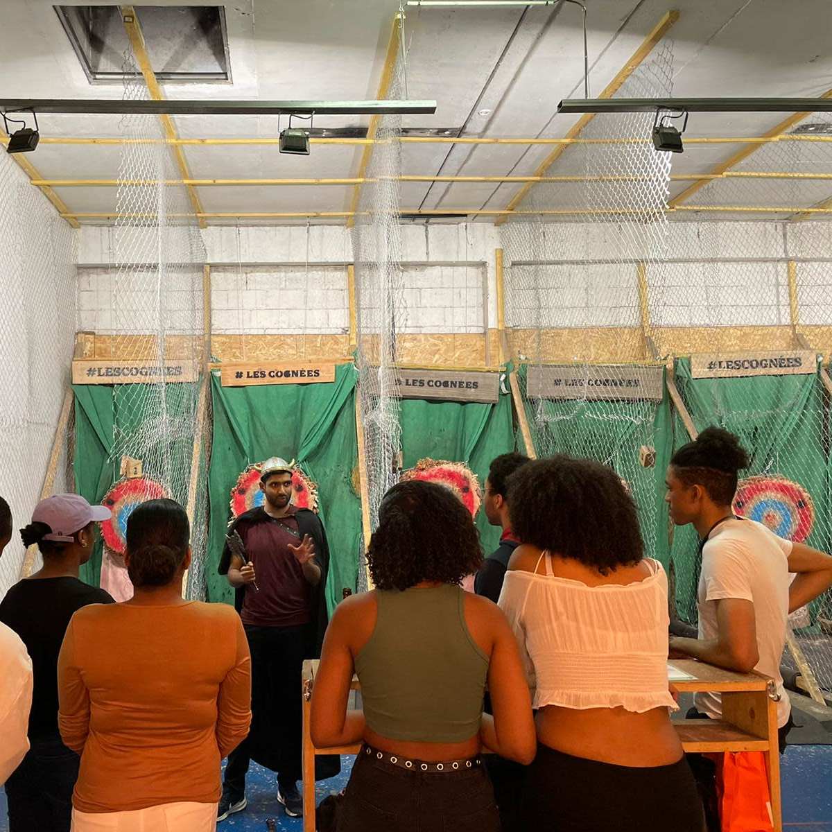A group of people listens to a speaker in an indoor axe-throwing venue surrounded by targets and safety nets.
