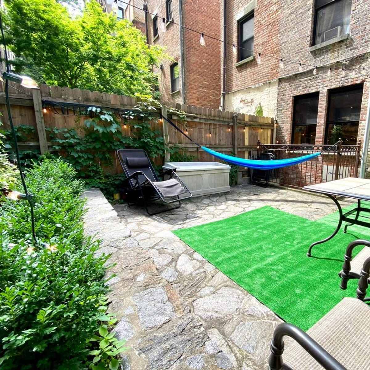 A small outdoor patio area with stone flooring, a green rug, a hammock, lounge chairs, and a table surrounded by greenery and brick walls.
