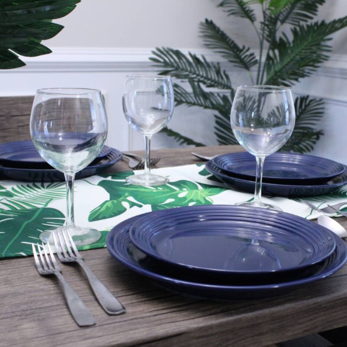 A dining table is set with navy plates, clear wine glasses, and silver cutlery, accompanied by a green leaf-patterned table runner and potted plants in the background.