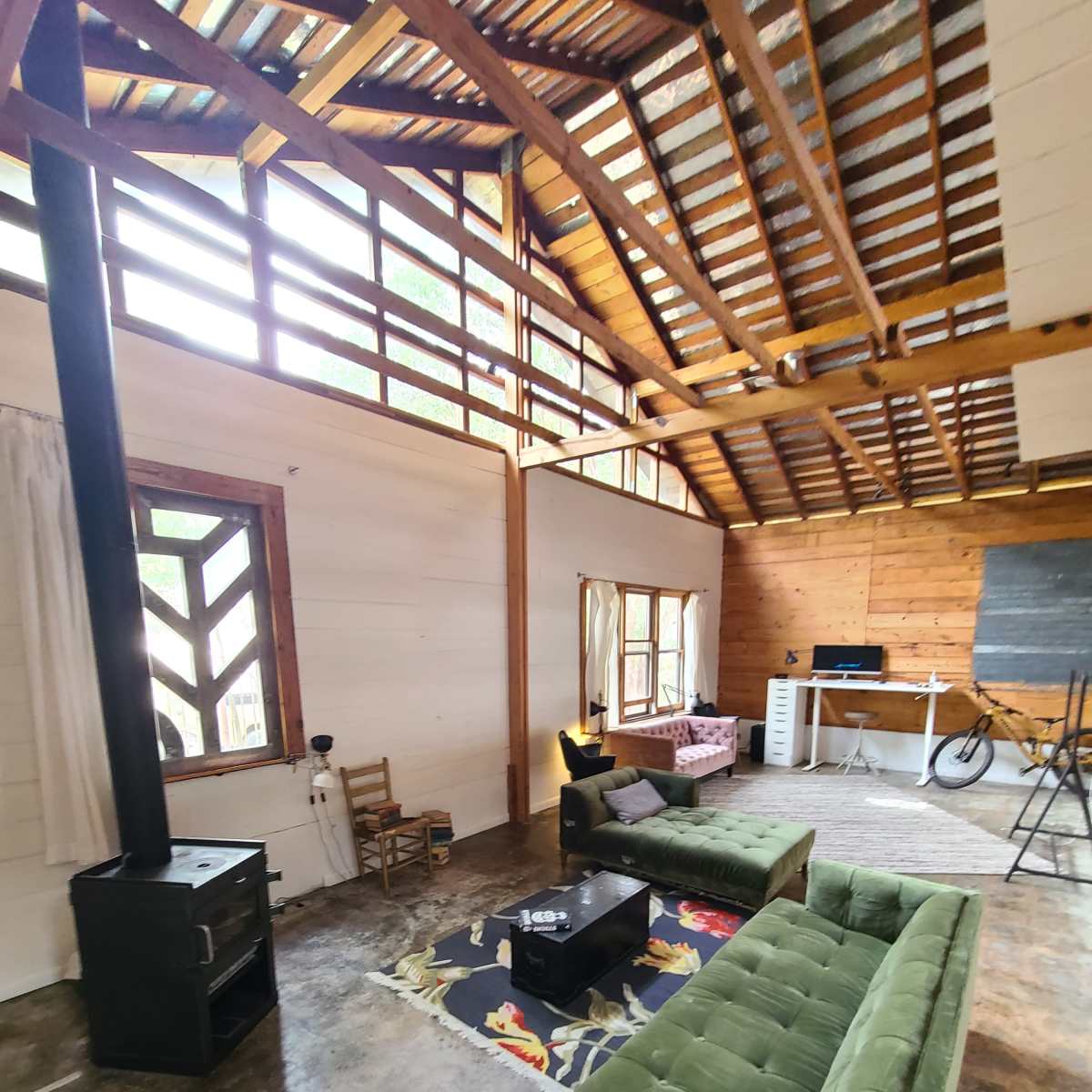 A spacious living area with exposed wooden beams, two green sofas, a black stove, a small table, and a workspace visible in the background.