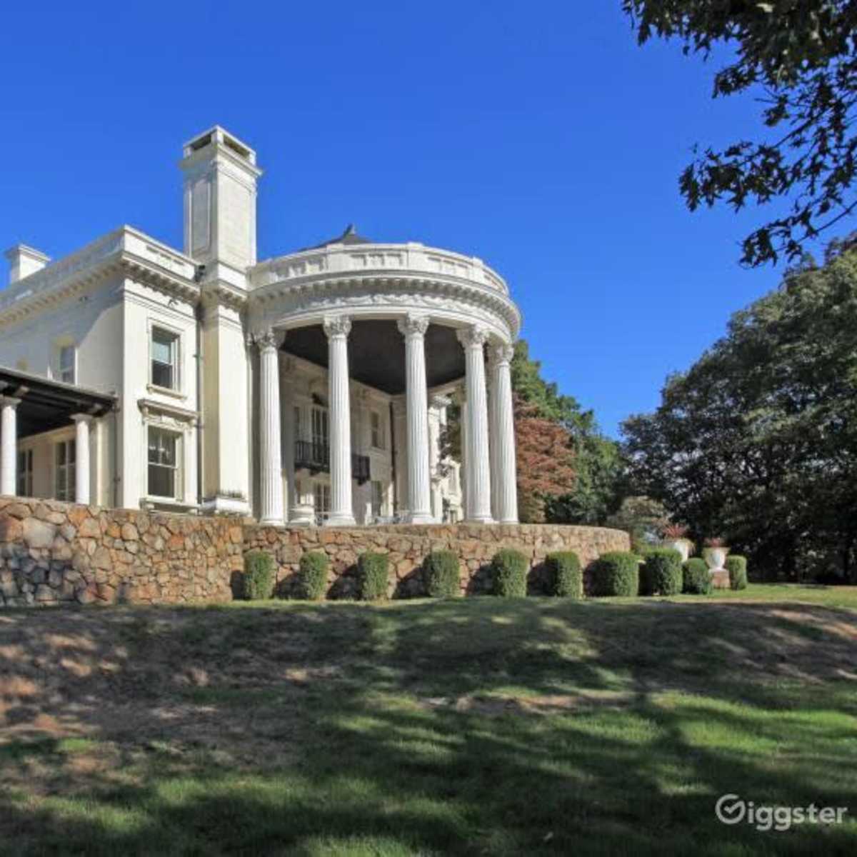 A large, white mansion with a circular portico sits atop a stone wall, surrounded by trees and grass under a clear blue sky.