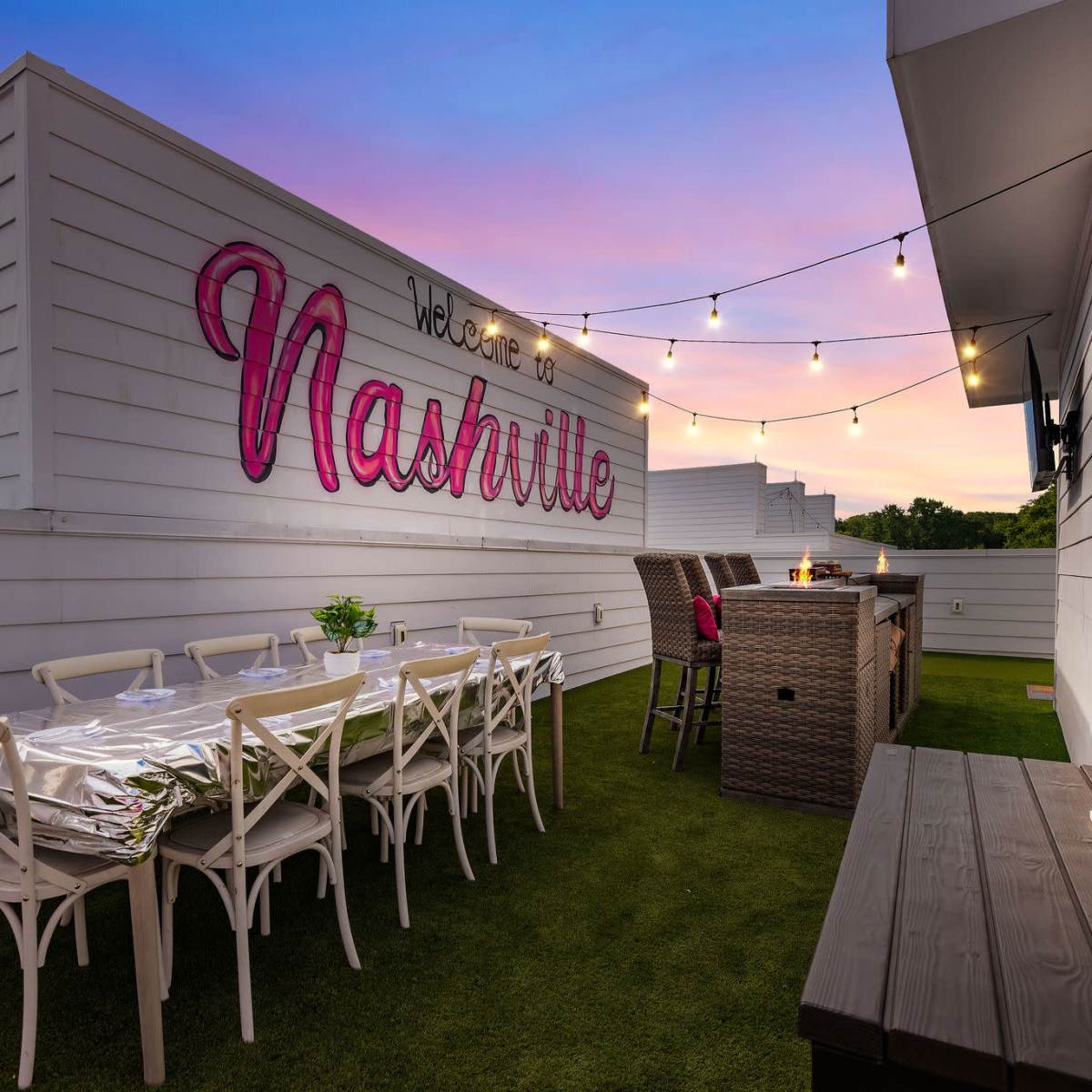A rooftop patio in Nashville adorned with string lights, a dining area with white chairs, and a bar setup against a backdrop of a colorful sunset.