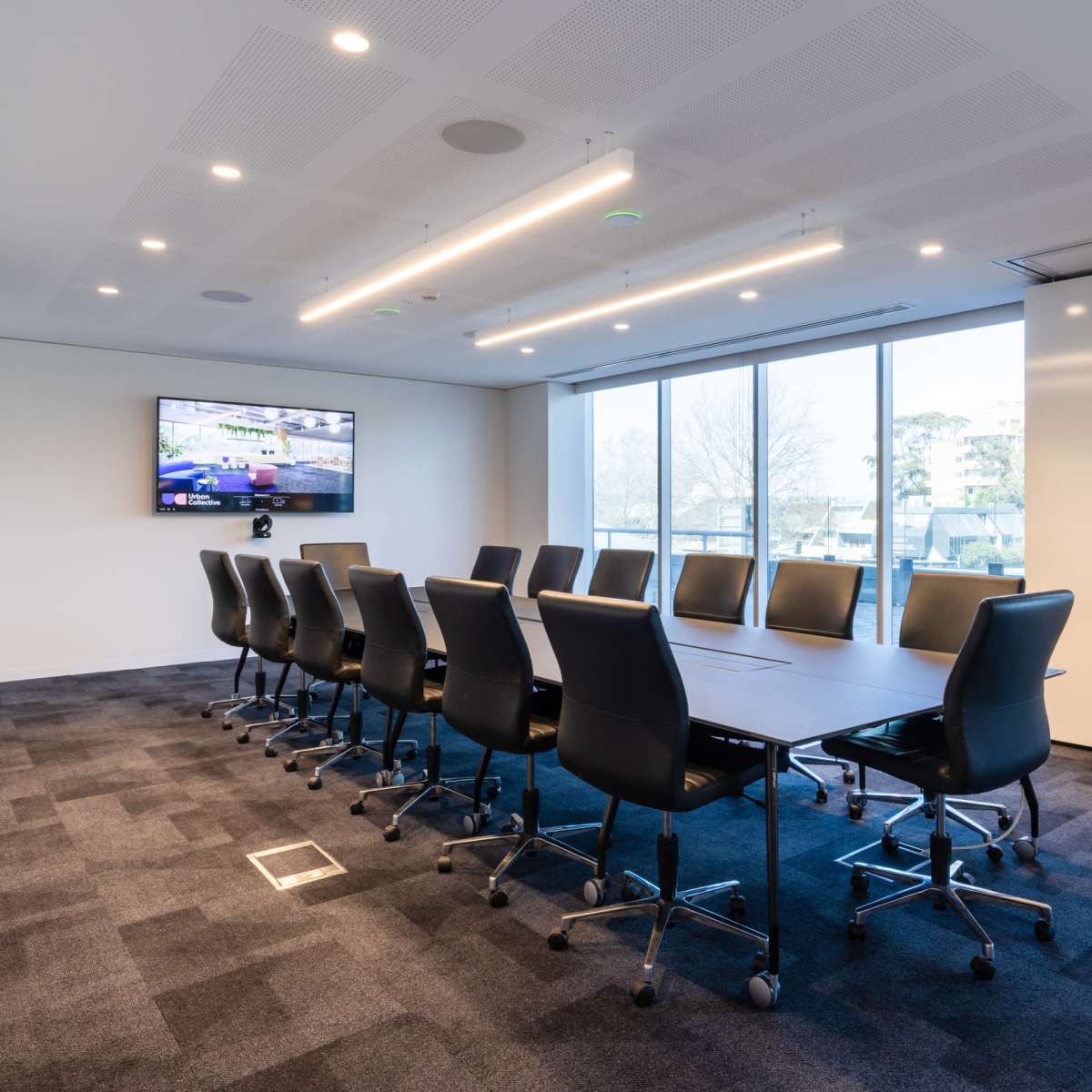 A modern conference room features a long table surrounded by black chairs, with a television mounted on the wall and large windows allowing natural light.