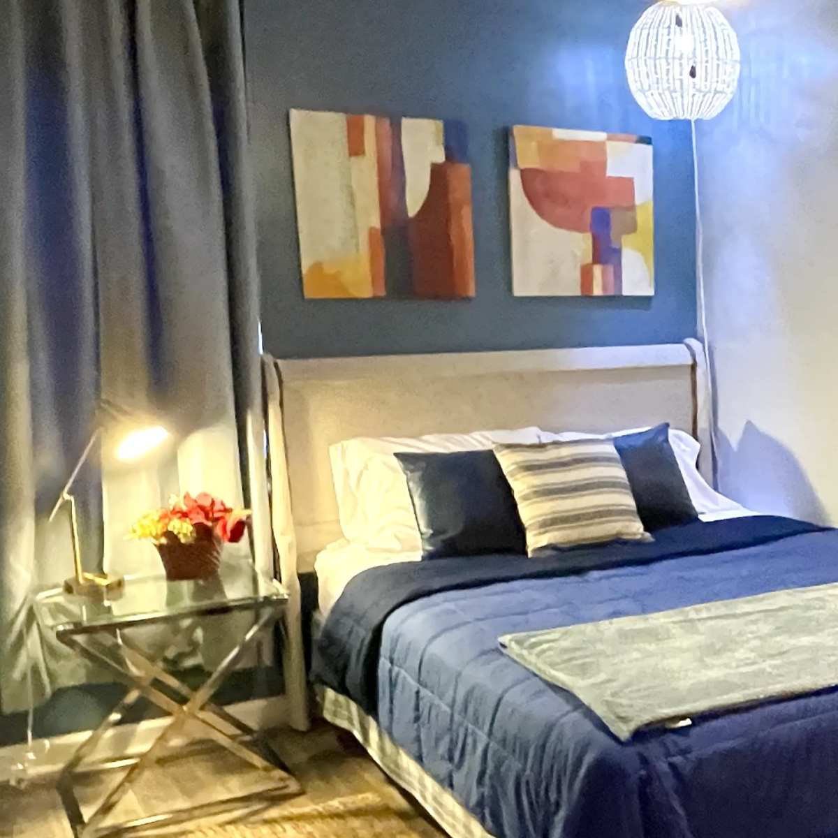 A neatly arranged bedroom with a bed adorned in blue bedding, a bedside table with a lamp, and two abstract paintings on the wall above the bed.